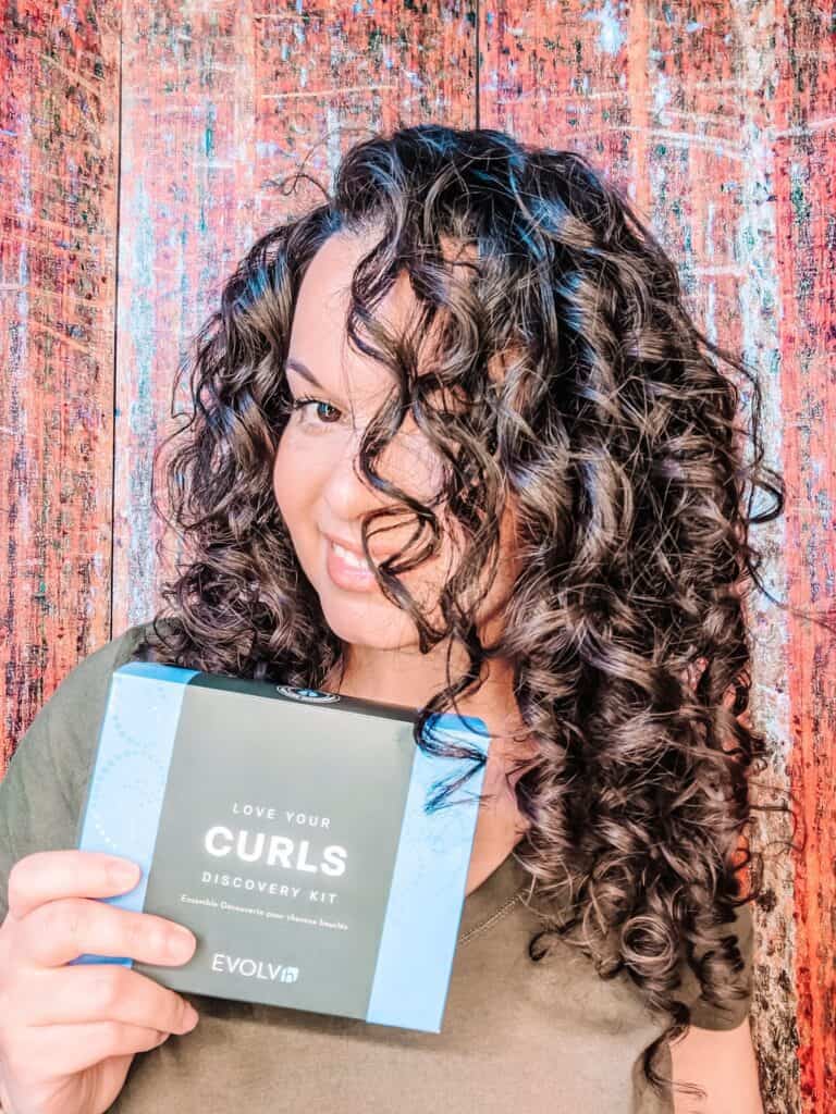 Delilah Orpi tilts head of 3a curly hair holding EVOLVh Curls discovery kit in front of reddish background